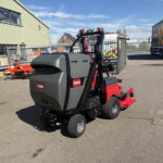 Industrial yard scene with a Toro ride-on attachment in the foreground, a green corrugated building on the left, and a large truck on the right.