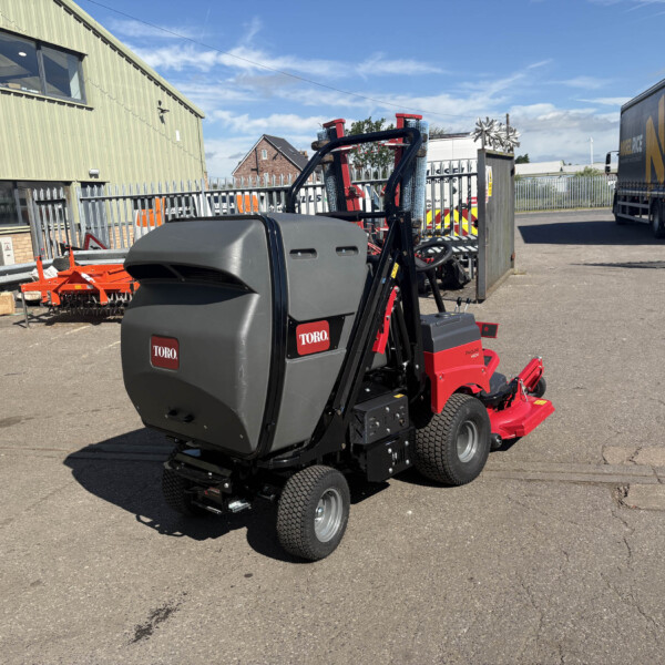 Industrial yard scene with a Toro ride-on attachment in the foreground, a green corrugated building on the left, and a large truck on the right.