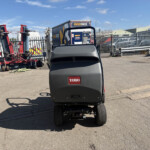 Rear view of a Toro ride-on mower parked in an industrial yard with red machinery and a blue trailer in the background