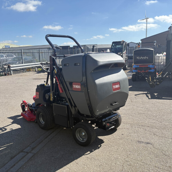 Close-up of a Toro riding lawn mower in an outdoor yard with metal fencing and various machinery in the background, including a Kubota machine farther back.
