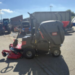 Red mower attachment hitched to a Kubota tractor in an outdoor equipment yard with other farm machinery and a wind turbine in the background on a sunny day.