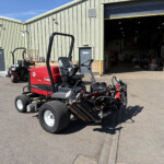 Red Toro Reelmaster ride-on mower parked outside a green metal warehouse with large open bay doors, equipment scattered nearby, bright sunny day.