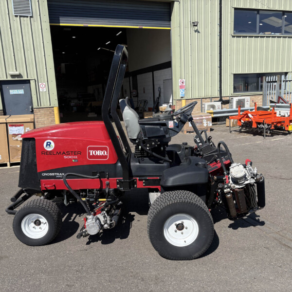 Red ride-on lawn mower parked outside a green industrial building with pallets and equipment nearby.
