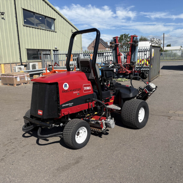 Red Toro Reel Master 5010-H riding mower in an outdoor yard next to a green metal building and a trailer, under a blue sky with scattered clouds.