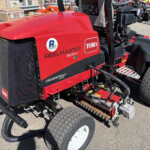 Red Toro Reelmaster 5010-H riding mower with large rear wheels and exposed cutting reels, parked outdoors with equipment nearby