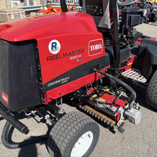 Red Toro Reelmaster 5010-H riding mower with large rear wheels and exposed cutting reels, parked outdoors with equipment nearby