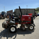 Red Toro Reelmaster 5010-D riding mower in a yard with industrial equipment and a blue sky in the background
