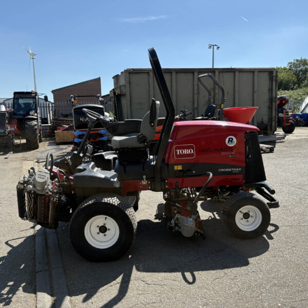 Red Toro Reelmaster 5010-D riding mower in a yard with industrial equipment and a blue sky in the background