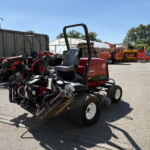 Outdoor yard with red farming tractors and pallets under a bright blue sky, a white greenhouse in the background.