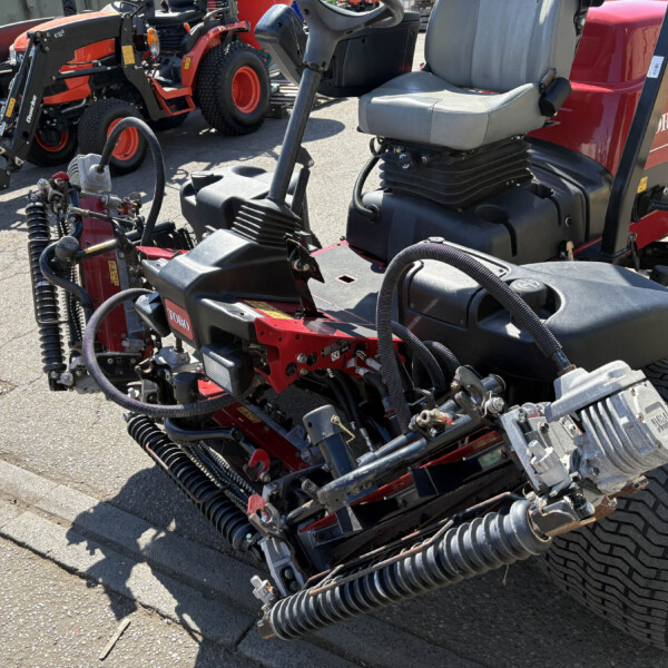 Close-up of a red Toro Reelmaster mowing unit showing hydraulic hoses, springs, and an elevated operator seat on a paved surface.