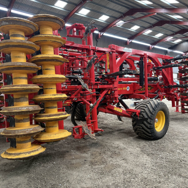 Red framed farming implement with large yellow disk harrows inside a metal workshop/warehouse.