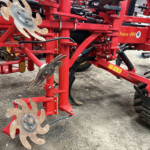 Red agricultural planter frame with rusted circular seed-discs and black hoses attached to a complex mechanism.