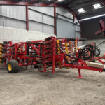 Red agricultural implement system with discs and tines parked inside a metal warehouse next to a tractor wheel.