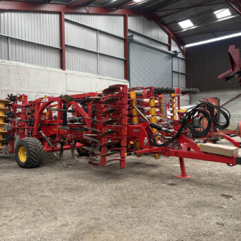 Red agricultural implement system with discs and tines parked inside a metal warehouse next to a tractor wheel.