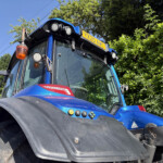 Blue tractor cab front with large tire in foreground, multiple lights, and a yellow plate reading YX20 AWC on top of the cab, against a hedge and blue sky.