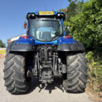 Front view of a large blue tractor with oversized tires, parked on a paved area beside a hedge and service buildings in the background.
