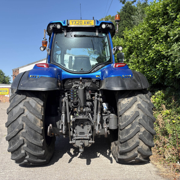 Front view of a large blue tractor with oversized tires, parked on a paved area beside a hedge and service buildings in the background.