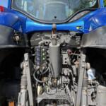 Close-up of a blue tractor's front-end hydraulics, levers, and control valves with exposed hoses and metal components.