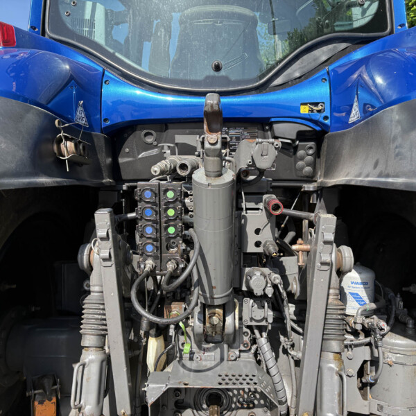 Close-up of a blue tractor's front-end hydraulics, levers, and control valves with exposed hoses and metal components.