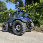 Blue farm tractor with oversized rear tires parked on a paved area beside a tall green hedge on a sunny day