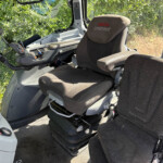 Interior of a Valtra Unlimited tractor cab with steering wheel, control console, and two dark fabric seats; plants visible through the window.
