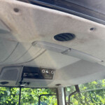 Interior of a car ceiling showing a round air vent, overhead panel and dashboard controls; outside greenery visible through windows.