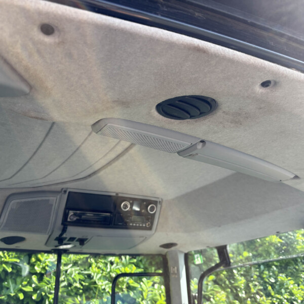 Interior of a car ceiling showing a round air vent, overhead panel and dashboard controls; outside greenery visible through windows.