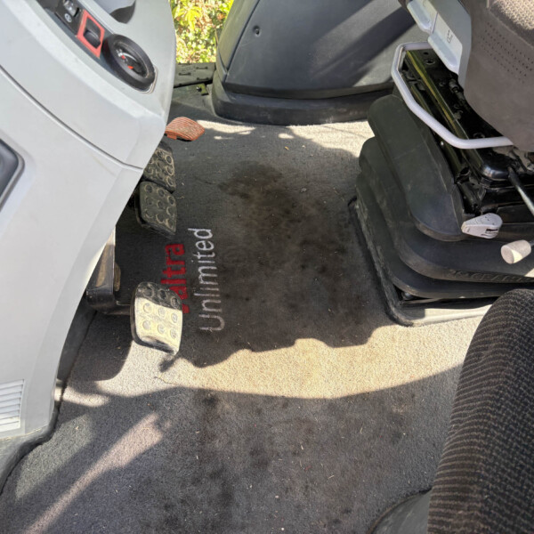 Driver’s footwell with two metal pedals and a black, dirty floor mat beside the seat