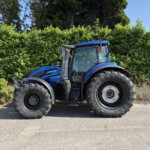 Blue tractor parked along a road beside a tall green hedge, clear blue sky above.
