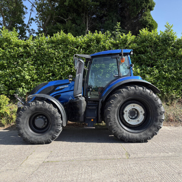 Blue tractor parked along a road beside a tall green hedge, clear blue sky above.