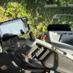 Interior of a heavy machinery cockpit with a joystick, multiple control panels, and a mounted screen; lush green foliage visible through the windshield