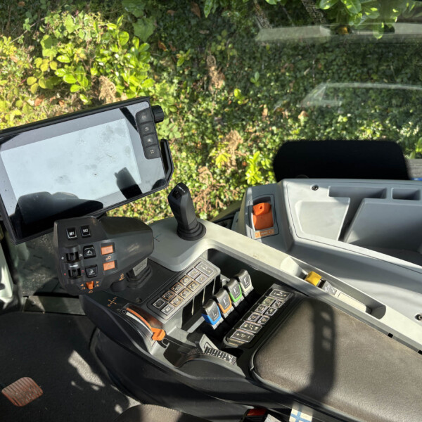 Interior of a heavy machinery cockpit with a joystick, multiple control panels, and a mounted screen; lush green foliage visible through the windshield
