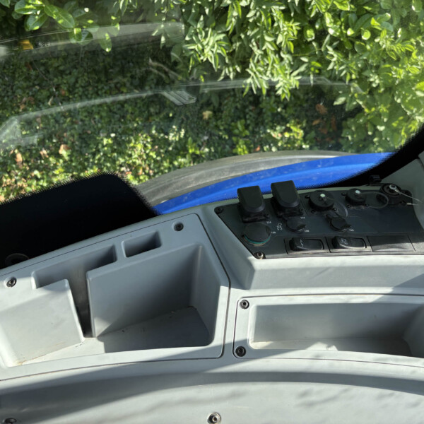 Vehicle cab interior with control panel and open storage compartments; windshield shows green hedge outside.