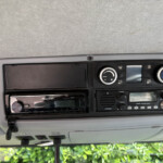 Dashboard center console with climate control knobs and a Pioneer stereo, in a grey interior with a windshield view of greenery outside.