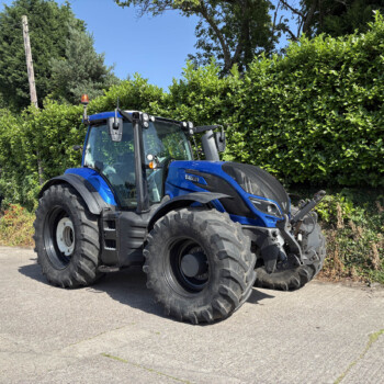 Blue agricultural tractor parked in a lot beside a tall green hedge on a sunny day, with trees in the background.