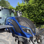 Close-up of a blue tractor cab with a large front tire, headlights, and a green hedge behind on a sunny day.