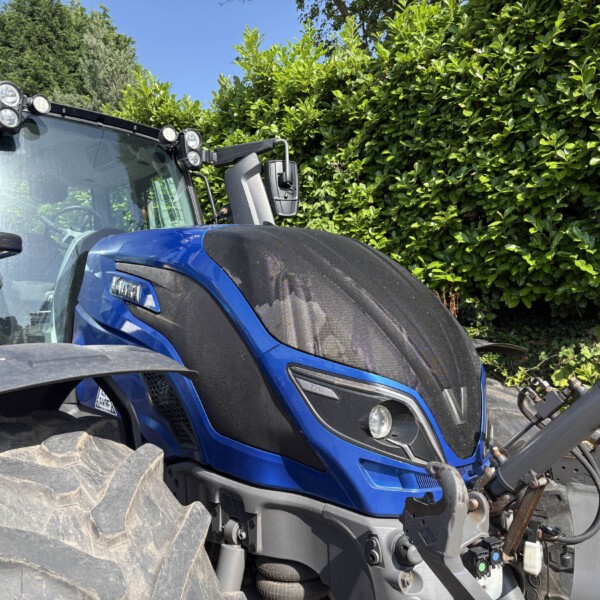 Close-up of a blue tractor cab with a large front tire, headlights, and a green hedge behind on a sunny day.