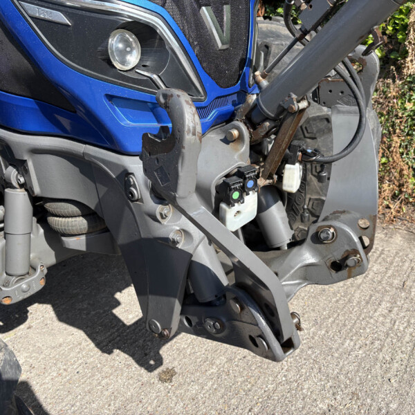 Close-up of a blue tractor front with hydraulic hitch, exposed metal arms, and hoses, parked on a paved surface with greenery in background