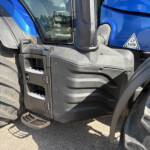 Close-up of a blue tractor cab showing the door area, black steps, and a large treaded tire with a dusty surface.