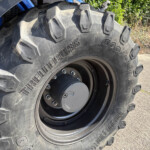 Close-up of a large off-road tractor tire with deep lug tread and a wheel hub, outdoors on pavement with a blue tractor body nearby (textured rubber visible).