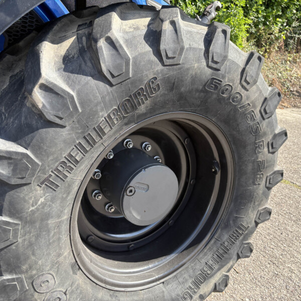 Close-up of a large off-road tractor tire with deep lug tread and a wheel hub, outdoors on pavement with a blue tractor body nearby (textured rubber visible).