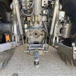 Underbelly view of heavy machinery showing hydraulic components, hoses, and metal linkages between two large tires.