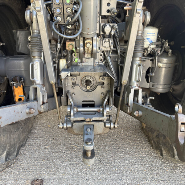 Underbelly view of heavy machinery showing hydraulic components, hoses, and metal linkages between two large tires.