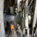 Close-up of a heavy machinery undercarriage showing a hydraulic hose, a bellows boot, and metal linkages near a large tire.