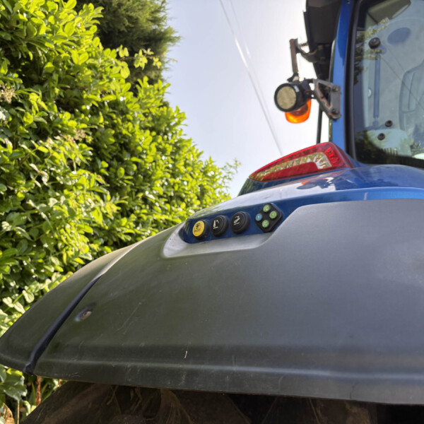 Blue utility vehicle with a round orange warning light mounted on a metal bracket, seen next to a dense green hedge and a dusty windshield edge.