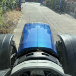 View from inside a blue tractor cab, the hood and dashboard visible as the vehicle travels down a narrow road flanked by hedges and a fence.