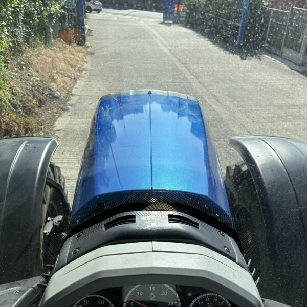 View from inside a blue tractor cab, the hood and dashboard visible as the vehicle travels down a narrow road flanked by hedges and a fence.