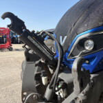 Close-up of a blue tractor's front hydraulic lift in the foreground, with red farming equipment and other tractors in the background.