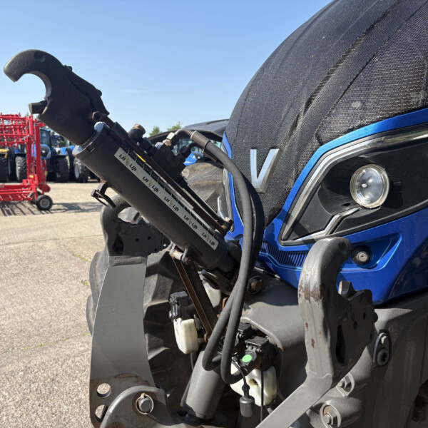 Close-up of a blue tractor's front hydraulic lift in the foreground, with red farming equipment and other tractors in the background.