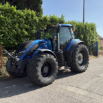 Blue tractor parked along a street beside a tall green hedge on a sunny day under a clear blue sky.
